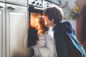 Little Toddler Girl And Two Kids Boys Baking, Pizza Or Cookies In Kitchen. Three Children, Siblings, Brothers And Sister Sitting Near Oven And Waiting. On Thanksgiving Or Christmas Holiday.